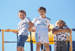 Drie lachende kinderen staan op een speeltoestel en genieten van een zonnige dag bij de lodge in de openlucht.