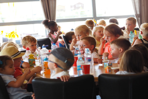 Kinderen zitten aan een tafel in een lodge, genieten van snacks, ijs en drankjes bij daglicht.