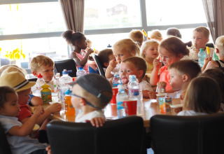 Kinderen zitten aan een tafel in een lodge, genieten van snacks, ijs en drankjes bij daglicht.
