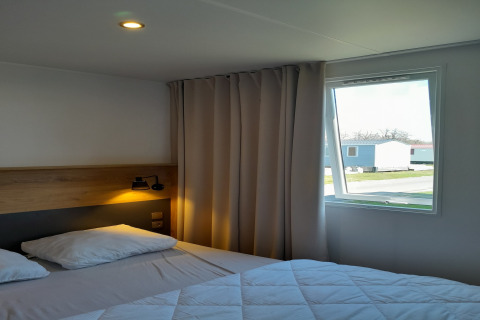 Camera moderna con letto matrimoniale, finestra aperta e vista su case mobili a Verblijfpark Breebos 4 in Belgio.