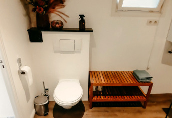 Baño moderno en Chalet «Chocolat» en Bélgica con inodoro, banco de madera, estantería y decoración floral.