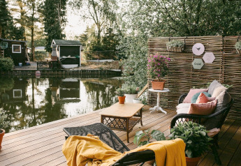 Acogedora terraza junto al agua en el Chalet Chocolat en Bélgica, con muebles de mimbre y plantas.