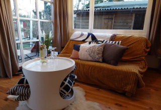 Acogedora sala en Casita La Luna, Bélgica, con sofá, cojines, mesa redonda y botella de vino.