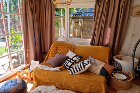 Acogedora sala en Casita La Luna, Bélgica, con sofá amarillo, cojines y ventanas grandes al jardín.