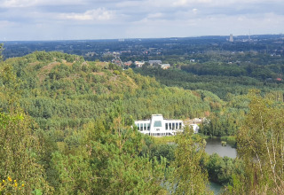 Vue sur un lodge moderne entouré de forêt dense, d'un étang, de collines et d'une ville au loin.