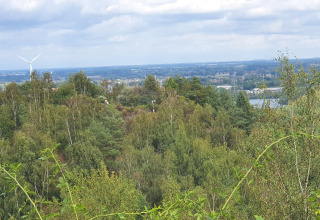Vue d'un lodge sur des collines verdoyantes, arbres, éolienne et ville lointaine sous ciel nuageux.