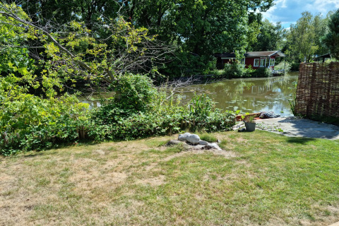Vista de un jardín verde junto al agua en el chalet Casita La Luna en Bélgica, con árboles y cabaña roja.