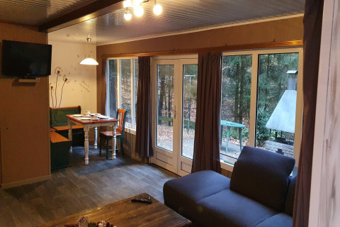 Sala en Forest House the Squirrel, Bélgica, con sofá, comedor, televisor y grandes ventanas al bosque.