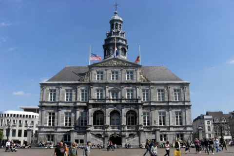 Des personnes marchent devant un hôtel de ville historique avec des drapeaux sur une place animée en Europe.