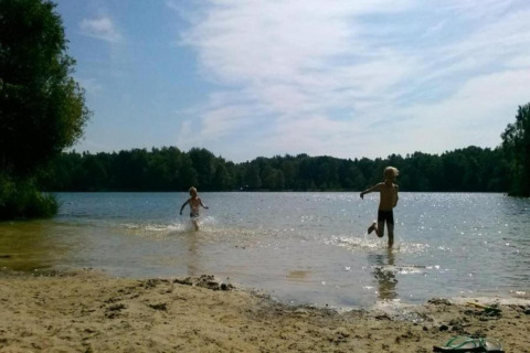 Two children play by the lakeshore under a sunny sky at a glamping accommodation surrounded by trees.