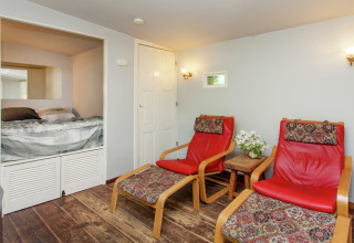 Knusse Vlaamse cabin in België, met twee rode stoelen, tafeltje met bloemen en hoekbed in een nis.