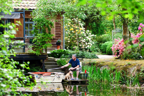 Man vist met hond aan een vijver voor een chalet, omringd door bloemen in een tuin in België.