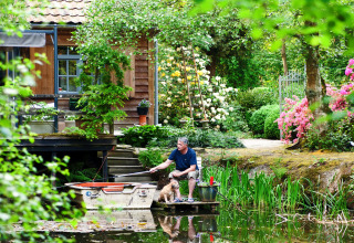 Man vist met hond aan een vijver voor een chalet, omringd door bloemen in een tuin in België.