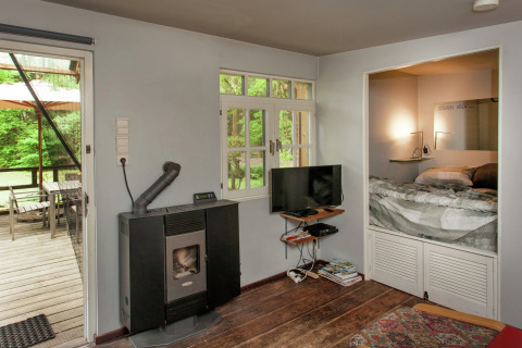 Gezellige woonkamer van The Wijerschans-cabine in België, met kachel, tv, nisbed en terraszicht.