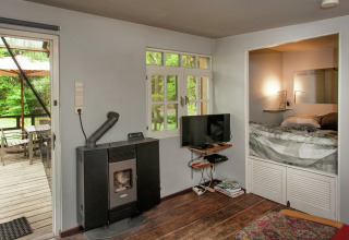 Gezellige woonkamer van The Wijerschans-cabine in België, met kachel, tv, nisbed en terraszicht.