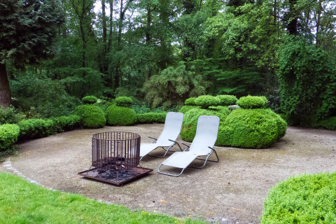 Twee ligstoelen en een vuurkorf op het terras van The Wijerschans-huisje in België, omringd door groen.