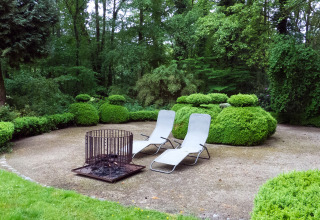 Twee ligstoelen en een vuurkorf op het terras van The Wijerschans-huisje in België, omringd door groen.