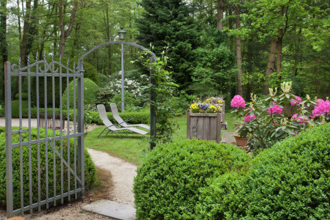 Tuin van ‘The Wijerschans’ in België met open ijzeren hek, ligstoelen, struiken en bloemen.
