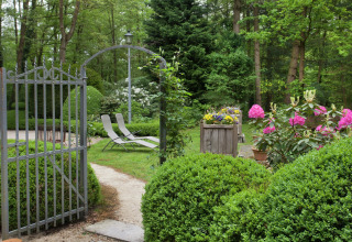 Tuin van ‘The Wijerschans’ in België met open ijzeren hek, ligstoelen, struiken en bloemen.
