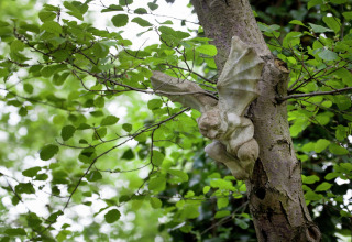 Stenen gargouille met vleugels bevestigd aan een boom tussen groene bladeren nabij The Wijerschans in België.