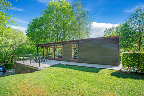 Lodge Manonfa moderno con grande terrazza, immerso nel verde degli alberi e sotto un cielo azzurro luminoso.