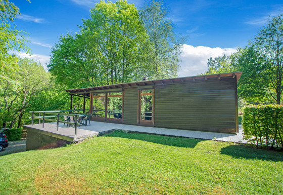 Lodge Manonfa moderno con grande terrazza, immerso nel verde degli alberi e sotto un cielo azzurro luminoso.