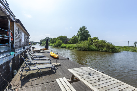 Solrige liggestole og bord på en husbådterrasse ved floden på St Antoine Isera i Belgien, omgivet af natur
