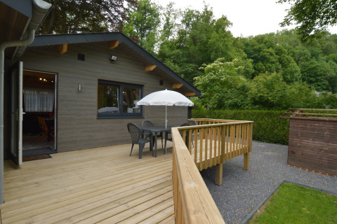 Wooden terrace at a chalet with table, chairs, and umbrella, surrounded by greenery in Belgium.