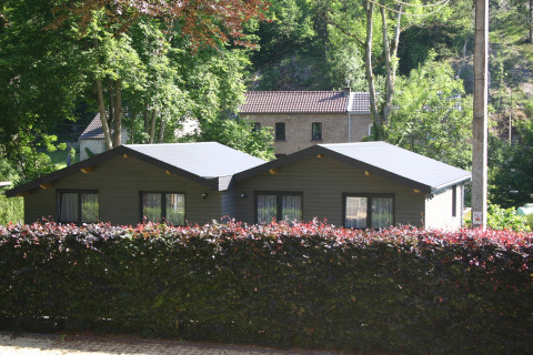 Glamping cabin with dark roof and large windows, surrounded by green trees and a hedge in nature.