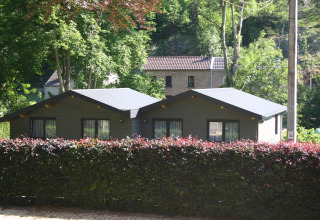 Glamping cabin with dark roof and large windows, surrounded by green trees and a hedge in nature.