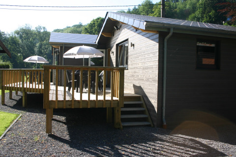 Le Chalet Chaleureux A, a wooden chalet with a deck, patio furniture and umbrellas, surrounded by nature.