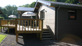 Le Chalet Chaleureux A, a wooden chalet with a deck, patio furniture and umbrellas, surrounded by nature.