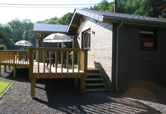 Le Chalet Chaleureux A, a wooden chalet with a deck, patio furniture and umbrellas, surrounded by nature.