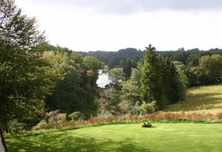 View from Le Chalet Chaleureux A in Belgium, showing a green lawn, trees, and a distant lake.