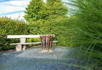 Vista del giardino verde allo Chalet «Orange» in Belgio con panchina di legno, ghiaia e cestino.