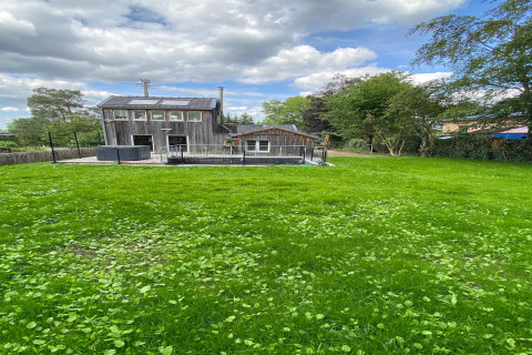 Foto van lodge 'Le coeur du brâme' in België met uitgestrekte grasveld, bomen en een bewolkte lucht erboven.