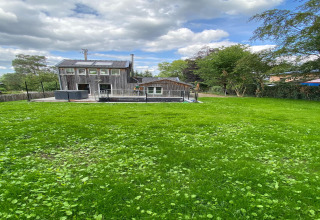 Foto van lodge 'Le coeur du brâme' in België met uitgestrekte grasveld, bomen en een bewolkte lucht erboven.