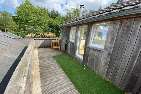 Houten terras met kunstgras en hoge stoelen bij een lodge in België, omringd door bomen en lucht.