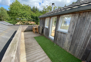 Houten terras met kunstgras en hoge stoelen bij een lodge in België, omringd door bomen en lucht.