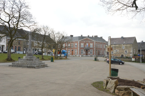 Rustig dorpsplein in België met een stenen monument, historische huizen en het gemeentehuis op de achtergrond.