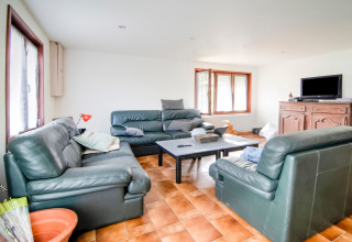 Living room with green leather sofas, coffee table, TV on wooden cabinet and large windows at a Belgian lodge.