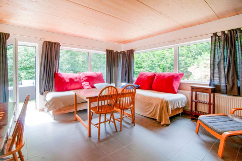 Bright lounge at Au Coeur du Pêcheur lodge in Belgium with big windows, wooden chairs, and forest view.