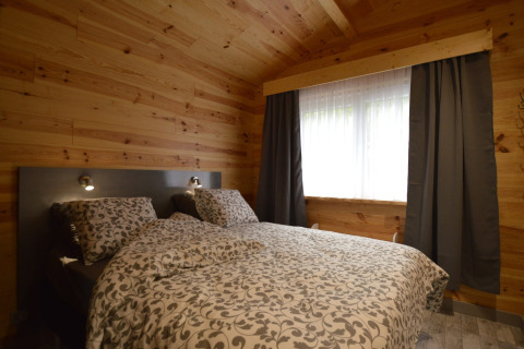 Gemütliches Schlafzimmer in Le Chalet Chaleureux B, Belgien, mit Holzwänden, Doppelbett und einem großen Fenster mit Vorhängen.