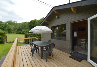 Holzterrasse mit Tisch, vier Stühlen und Sonnenschirm beim Le Chalet Chaleureux B, umgeben von Natur in Belgien.
