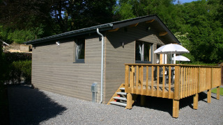 Exterior of Le Chalet Chaleureux B, a wooden lodge featuring a deck with umbrellas, set in lush greenery.