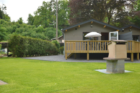 Le Chalet Chaleureux B in Belgien mit Holzterrasse, Sonnenschirm, Rasen und gemauertem Grillplatz.