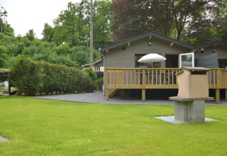 Le Chalet Chaleureux B in Belgien mit Holzterrasse, Sonnenschirm, Rasen und gemauertem Grillplatz.