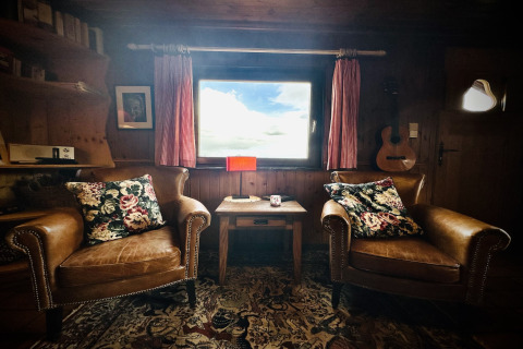 Gezellig interieur van La Cabane in België met leren stoelen, bloemenkussens, gitaar en uitzicht naar buiten.