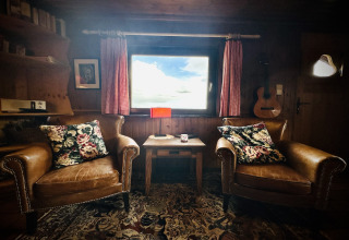 Gezellig interieur van La Cabane in België met leren stoelen, bloemenkussens, gitaar en uitzicht naar buiten.