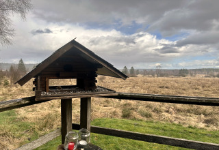 Mangiatore per uccelli su terrazza in legno a La Cabane, Belgio, affacciato su un campo aperto e cielo nuvoloso.